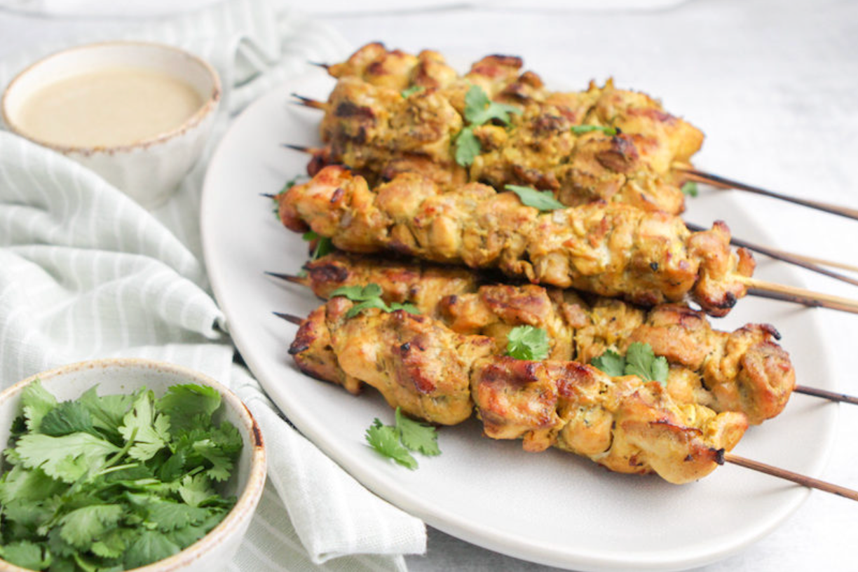 Skewers of chicken satay with a side of cilantro on a white plate.